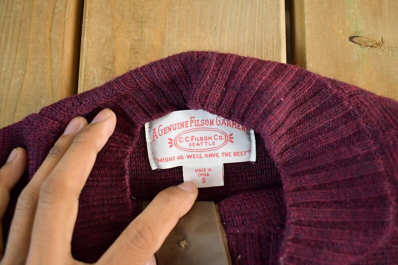Puede incluir: Primer plano de un cuello de su&eacute;ter burdeos con textura acanalada. Una etiqueta blanca muestra el texto "A Genuine Filson Garment" y "Might as well have the best". La etiqueta tambi&eacute;n indica la talla S.