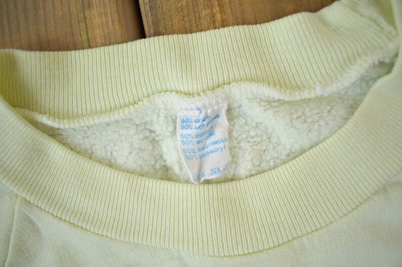 May include: A pale yellow sweatshirt with a blue and white care label that reads "60% cotton 40% acrylic 50% cotton 50% wool 50% polyester".