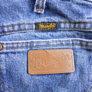 May include: Close-up of a pair of blue denim jeans with a brown leather Wrangler label on the back pocket. The label has the word "Wrangler" stitched in white thread.