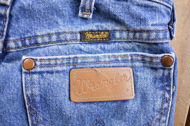 May include: Close-up of a pair of blue denim jeans with a brown leather Wrangler label on the back pocket. The label has the word "Wrangler" stitched in white thread.