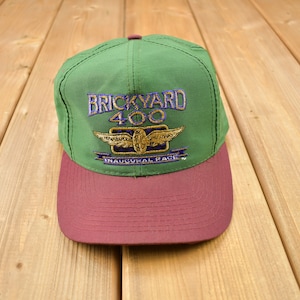 May include: Green and maroon baseball cap with the text "Brickyard 400" and a logo of a plane with a chequered flag. The text "Inaugural Race" is below the logo.