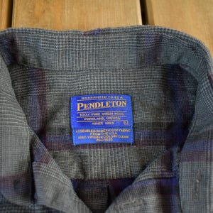 Puede incluir: Primer plano de una camisa abotonada a cuadros gris, azul y burdeos. La camisa tiene cuello y una etiqueta azul que dice "PENDLETON 100% PURE VIRGIN WOOL PORTLAND, OREGON SINCE 1963". La camisa est&aacute; hecha de una tela suave y tejida.