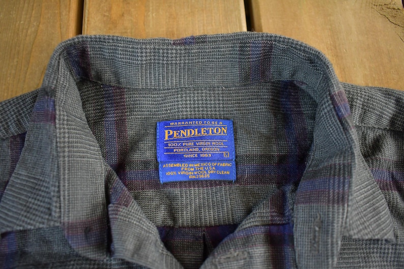 Puede incluir: Primer plano de una camisa abotonada a cuadros gris, azul y burdeos. La camisa tiene cuello y una etiqueta azul que dice "PENDLETON 100% PURE VIRGIN WOOL PORTLAND, OREGON SINCE 1963". La camisa est&aacute; hecha de una tela suave y tejida.