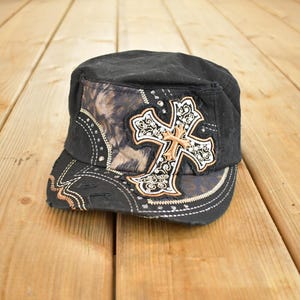 Può includere: Cappellino nero con un design a croce decorativo. Il cappellino presenta un motivo mimetico sul lato e una grande croce ornata con accenti di strass. L'orlo e i bordi hanno cuciture decorative.