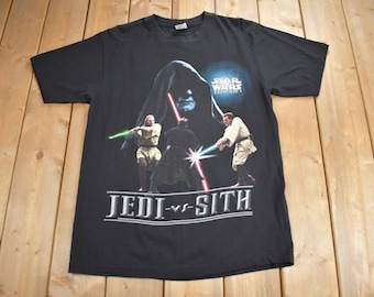 Könnte beinhalten: Schwarzes T-Shirt mit einem Aufdruck von drei Star Wars-Charakteren, einem Jedi und einem Sith, mit Lichtschwertern. Der Text "Star Wars Episode I" und "Jedi vs Sith" ist auf dem T-Shirt aufgedruckt.