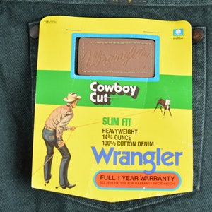 May include: A pair of dark green Wrangler jeans with a yellow and green tag attached. The tag features a cowboy riding a horse, the Wrangler logo, and the text "Cowboy Cut", "Slim Fit", "Heavyweight 14 1/4 ounce", "100% Cotton Denim", and "Full 1 Year Warranty".