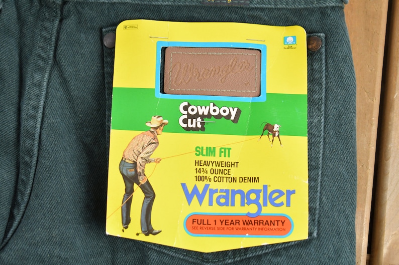 May include: A pair of dark green Wrangler jeans with a yellow and green tag attached. The tag features a cowboy riding a horse, the Wrangler logo, and the text "Cowboy Cut", "Slim Fit", "Heavyweight 14 1/4 ounce", "100% Cotton Denim", and "Full 1 Year Warranty".