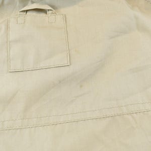 May include: Close-up of a tan jacket with a square pocket and a small flap. The jacket has visible stitching and a slightly wrinkled texture. The fabric appears to be lightweight.