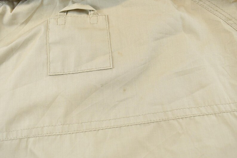 May include: Close-up of a tan jacket with a square pocket and a small flap. The jacket has visible stitching and a slightly wrinkled texture. The fabric appears to be lightweight.
