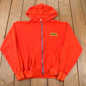 Peut inclure: Un sweat-shirt à capuche rouge avec une fermeture éclair et un patch jaune avec le mot "KENT" sur le devant.