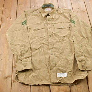 Peut inclure: Une chemise à manches longues de couleur beige avec deux bandes vertes sur chaque manche. La chemise a deux grandes poches sur le devant et une fermeture à boutons.