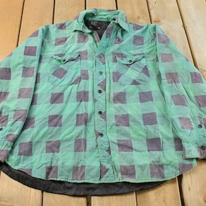 May include: A green and purple checked button-down shirt with two chest pockets. The shirt has long sleeves and a black lining at the bottom. The shirt is laid out on a wooden surface.