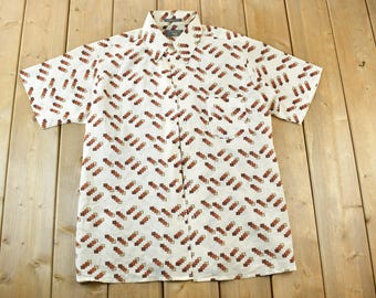May include: White short-sleeve button-down shirt with a brown geometric pattern. The shirt has a collared neckline, a chest pocket, and a button-up front. The pattern is repeated across the shirt's surface.