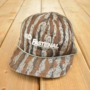 Può includere: Un cappellino da camionista Fastenal vintage con un motivo mimetico marrone, grigio e bianco. Il cappello presenta il logo Fastenal in bianco sul davanti. Il cappello ha un retro in rete e una visiera curva. Un dettaglio in corda verde e bianca è sul bordo.