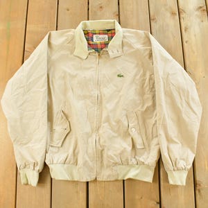 May include: Tan zip-up jacket with a light-colored collar and cuffs. The jacket features two front pockets and a small green crocodile logo on the chest. The interior collar lining has a plaid pattern.