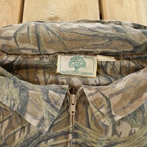 Op de afbeelding: Close-up van een camouflage shirt met ritssluiting. Het shirt heeft een bruin, groen en beige patroon. De kraag is omgeslagen en een label met het "Mossy Oak" logo is zichtbaar. De rits is beige.