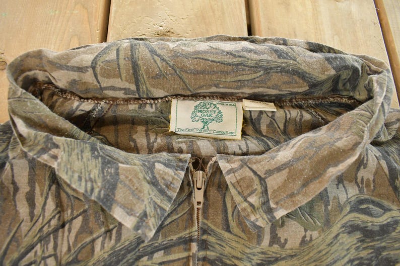 Op de afbeelding: Close-up van een camouflage shirt met ritssluiting. Het shirt heeft een bruin, groen en beige patroon. De kraag is omgeslagen en een label met het "Mossy Oak" logo is zichtbaar. De rits is beige.