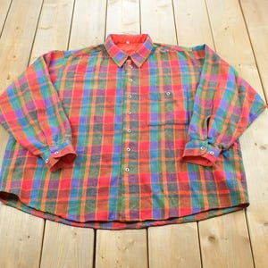 Peut inclure: Une chemise à manches longues boutonnée avec un motif à carreaux vif dans des tons de rouge, bleu, vert et orange. La chemise comprend un col classique, une poche poitrine unique sur le côté gauche et des poignets boutonnés sur chaque manche.