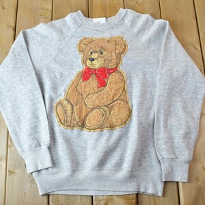 May include: A gray sweatshirt with a brown teddy bear wearing a red bow tie. The bear is embroidered on the front of the sweatshirt.