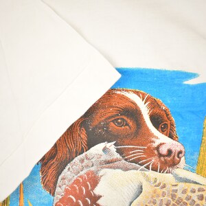 May include: A brown and white dog with a duck in its mouth. The dog is looking at the viewer. The background is a blue sky with green reeds.