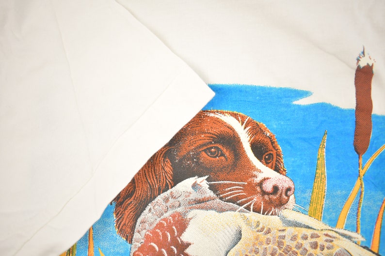 May include: A brown and white dog with a duck in its mouth. The dog is looking at the viewer. The background is a blue sky with green reeds.
