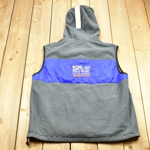 Puede incluir: Chaleco de forro polar gris con una franja azul y naranja en el pecho. El chaleco tiene capucha y el texto "PRL-67 POLO SPORT RALPH LAUREN MARINE SYSTEM" está impreso en la franja azul.