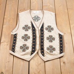 Op de afbeelding: Een crèmekleurige gebreide vest met een bruin en groen geometrisch patroon. De vest heeft een knoopsluiting aan de voorkant en een V-hals.