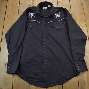 Puede incluir: Una camisa negra de manga larga con botones, con bordados blancos de plumas y un diseño de calavera. La camisa tiene dos bolsillos en el pecho y un cuello puntiagudo.