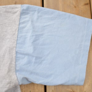 May include: Close-up of the sleeve of a two-tone t-shirt. The sleeve is light blue and the body of the shirt is a light gray.