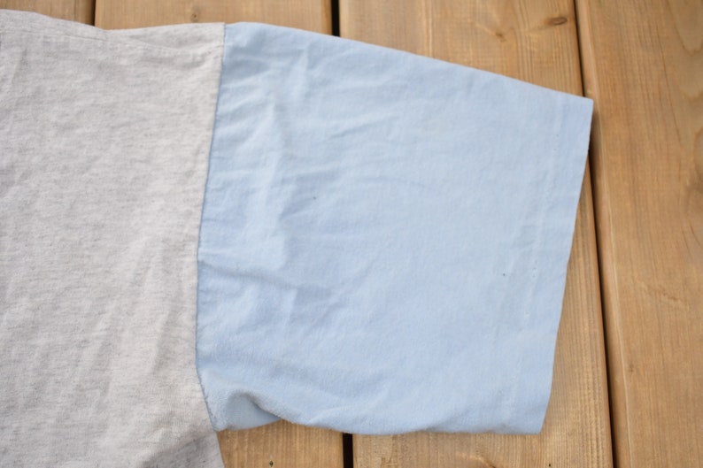 May include: Close-up of the sleeve of a two-tone t-shirt. The sleeve is light blue and the body of the shirt is a light gray.