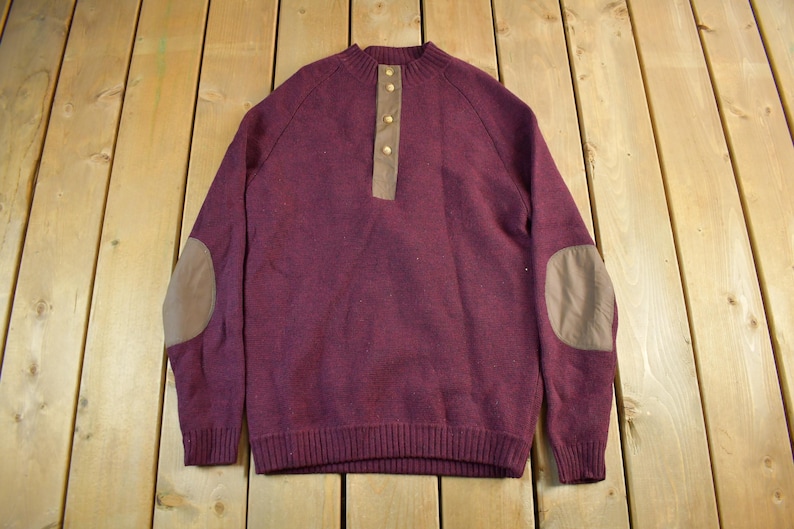 Puede incluir: Un su&eacute;ter burdeos con dobladillo y pu&ntilde;os acanalados. El su&eacute;ter presenta una tapeta abotonada, coderas y cuello redondo. Las coderas y la tapeta son de color marr&oacute;n claro. El su&eacute;ter est&aacute; hecho de un material suave y c&aacute;lido.