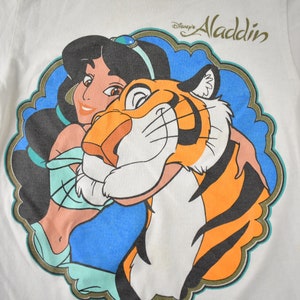 May include: White t-shirt with a colorful graphic of Jasmine from Disney's Aladdin holding Rajah the tiger. The text "Disney's Aladdin" is printed in gold above the graphic.