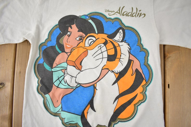 May include: White t-shirt with a colorful graphic of Jasmine from Disney's Aladdin holding Rajah the tiger. The text "Disney's Aladdin" is printed in gold above the graphic.