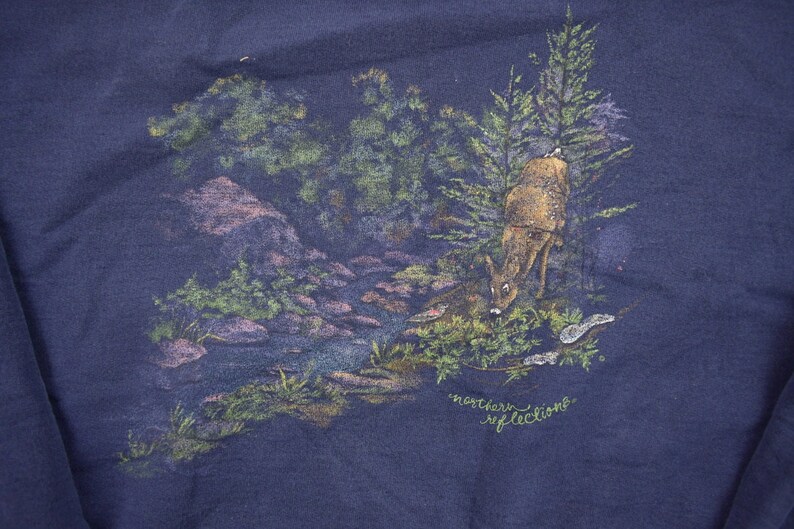 Puede incluir: Una sudadera azul marino con una escena de naturaleza impresa en la parte delantera. La escena presenta un ciervo bebiendo de un arroyo en un entorno de bosque. El texto "northern reflections" est&aacute; impreso debajo de la escena.