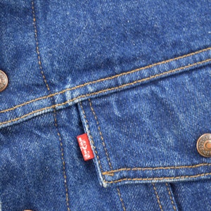 May include: Close-up of a blue denim jacket with a red Levi's patch on the pocket. The jacket has two copper-colored buttons and a brown stitching detail.