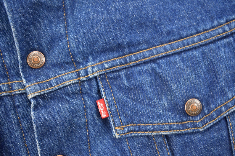 May include: Close-up of a blue denim jacket with a red Levi's patch on the pocket. The jacket has two copper-colored buttons and a brown stitching detail.
