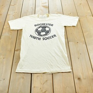 May include: A cream-colored short-sleeve t-shirt featuring a black graphic of a soccer ball in the center. The text "ROCHESTER" is printed above the soccer ball, and the text "YOUTH SOCCER" is printed below the soccer ball in black lettering.