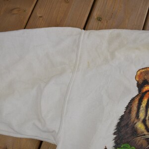 K&ouml;nnte beinhalten: Wei&szlig;er Pullover mit einem Tiger-Grafikdruck. Der Tiger hat orange und schwarze Streifen und schaut direkt auf den Betrachter.