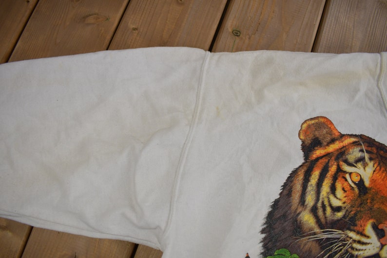 K&ouml;nnte beinhalten: Wei&szlig;er Pullover mit einem Tiger-Grafikdruck. Der Tiger hat orange und schwarze Streifen und schaut direkt auf den Betrachter.