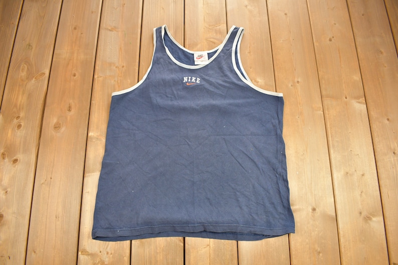 May include: A navy blue sleeveless tank top with white trim around the neck and armholes. The tank top has a white Nike logo and the word "NIKE" in red lettering on the front.
