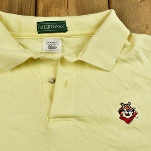 K&ouml;nnte beinhalten: Ein gelbes Poloshirt mit einem gr&uuml;nen "OUTER BANKS"-Etikett und einem gestickten Cartoon-Tiger in Rot und Blau, der ein rotes Bandana tr&auml;gt.