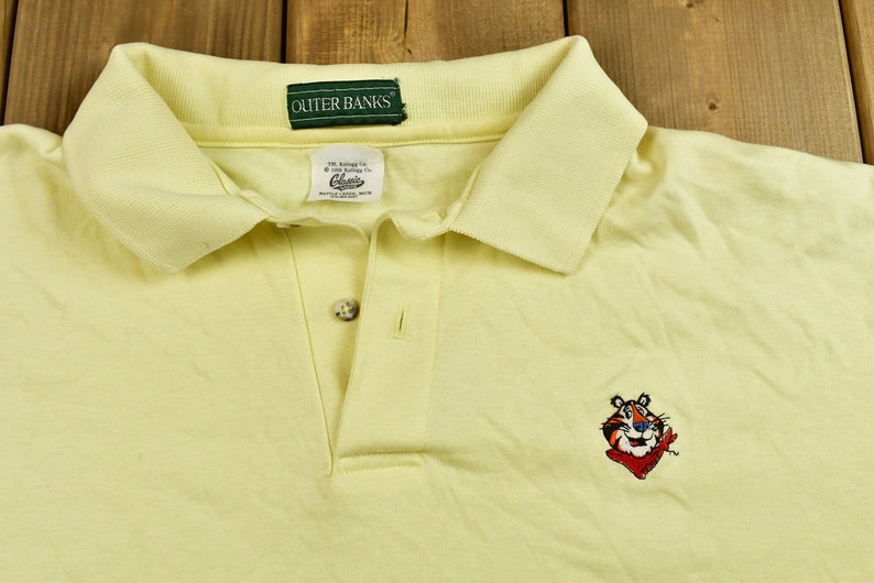 K&ouml;nnte beinhalten: Ein gelbes Poloshirt mit einem gr&uuml;nen "OUTER BANKS"-Etikett und einem gestickten Cartoon-Tiger in Rot und Blau, der ein rotes Bandana tr&auml;gt.