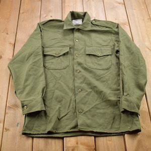 Puede incluir: Una camisa verde de manga larga con cierre de botones y dos bolsillos grandes. La camisa tiene cuello y puños con botones.