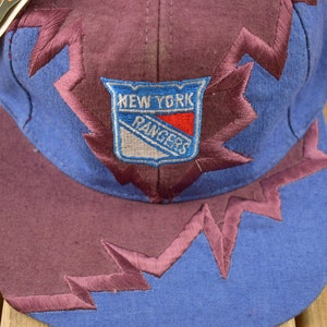 May include: A blue and purple baseball cap with a New York Rangers logo embroidered on the front. The cap has a jagged, lightning bolt design around the brim.