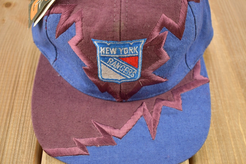 May include: A blue and purple baseball cap with a New York Rangers logo embroidered on the front. The cap has a jagged, lightning bolt design around the brim.