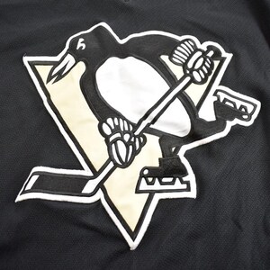 Puede incluir: Camiseta de hockey negra y dorada con un logotipo de ping&uuml;ino. El ping&uuml;ino sostiene un palo de hockey y est&aacute; de pie sobre un disco de hockey negro y blanco.