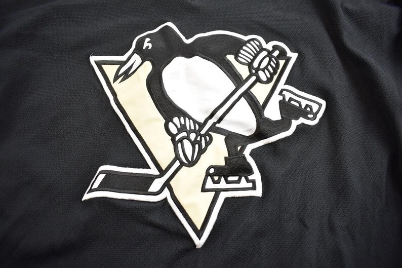 Puede incluir: Camiseta de hockey negra y dorada con un logotipo de ping&uuml;ino. El ping&uuml;ino sostiene un palo de hockey y est&aacute; de pie sobre un disco de hockey negro y blanco.