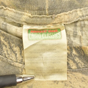 May include: Close-up of a camouflage pattern t-shirt with a tag that reads "Mossy Oak Camo Classics" in red and green. The shirt has a crew neck and is made of a soft, textured fabric. The camouflage design features shades of brown, green, and gray.