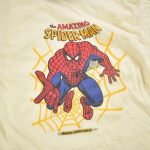 May include: A vintage white t-shirt with a cartoon image of Spider-Man in a red and blue suit, swinging on a web. The text "The Amazing Spider-Man" is printed above the image. The copyright information "&copy; MARVEL COMICS GROUP 1975" is printed below the image.