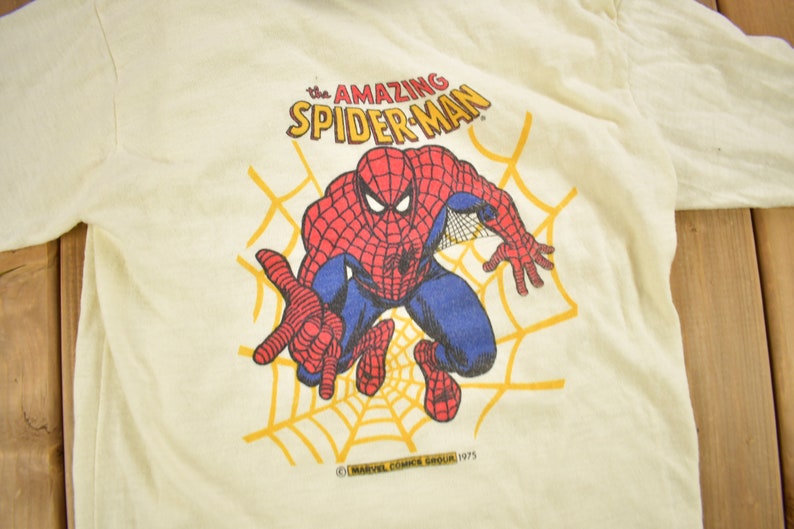 May include: A vintage white t-shirt with a cartoon image of Spider-Man in a red and blue suit, swinging on a web. The text "The Amazing Spider-Man" is printed above the image. The copyright information "&copy; MARVEL COMICS GROUP 1975" is printed below the image.
